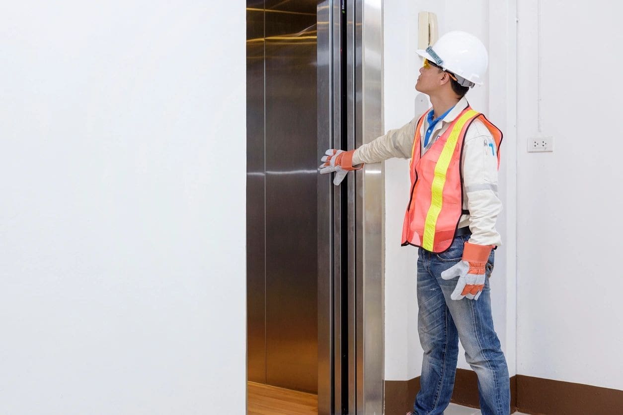 A man in an orange vest and hard hat is opening the door of a lift.